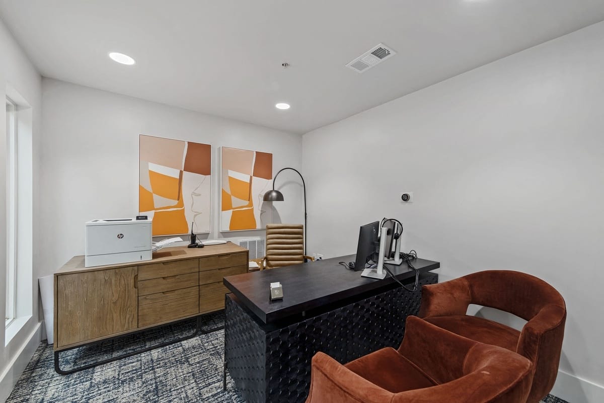 Business center with comfy chairs, a desk and printer on cabinet next to a lamp.