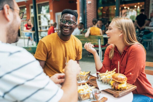 Three individuals enjoying a meal together at an outdoor restaurant, surrounded by greenery and a pleasant atmosphere.