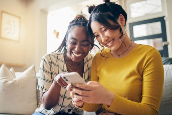 Two women on a couch, engaged with their phones, showcasing a moment of leisure and connection in a cozy setting.