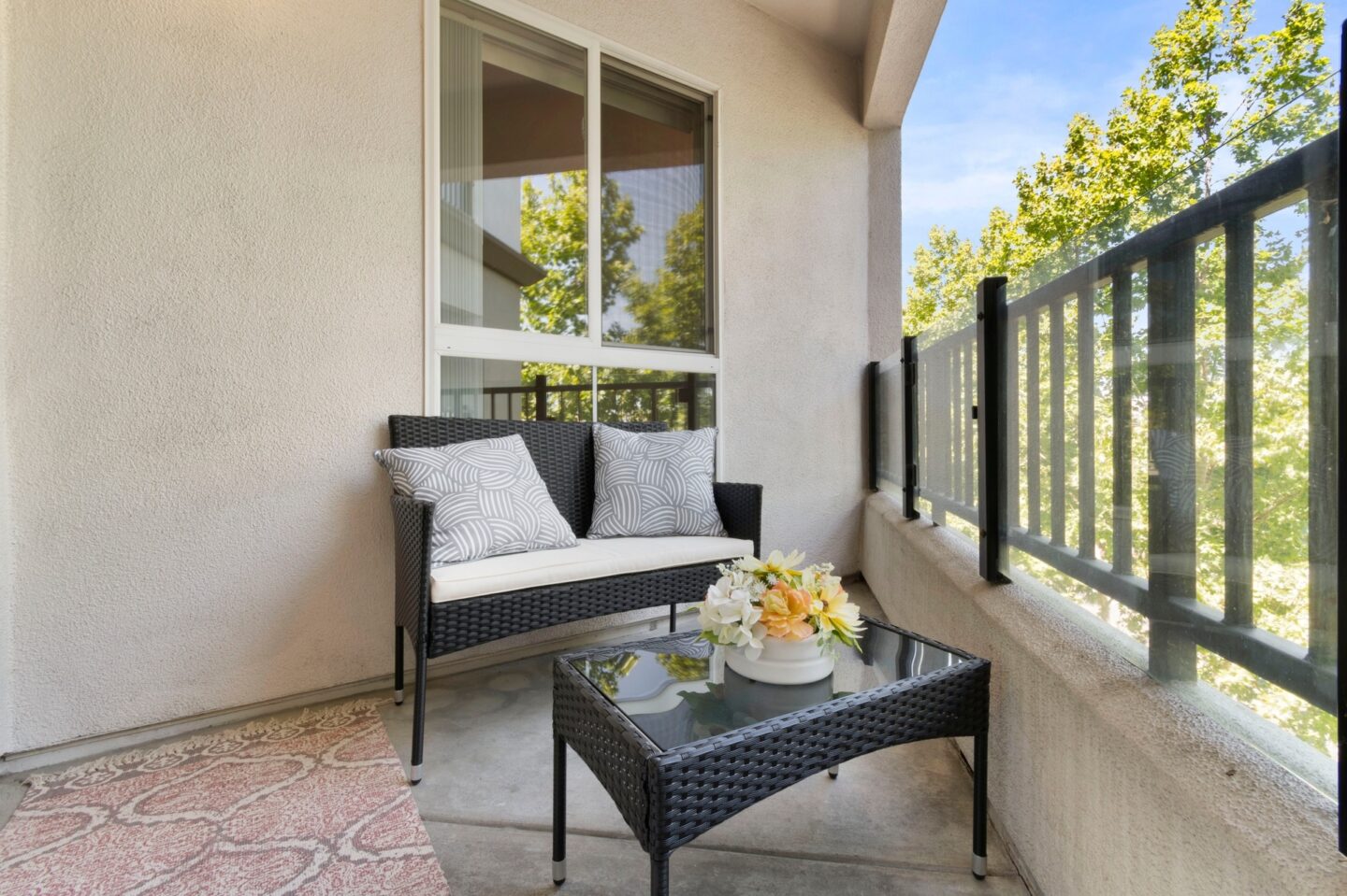 A patio with a black wicker sofa and a glass table with a vase of flowers on it.