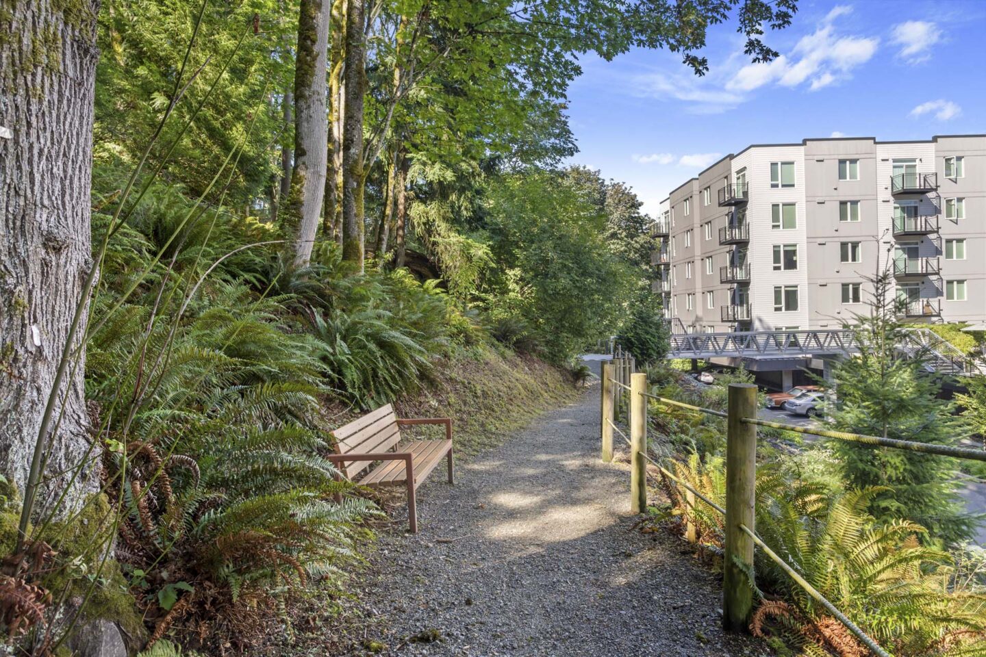 A gravel path in a wooded area with a bench and apartment building in the background at Windsor Totem Lake, 11723 NE 117th Ct, Kirkland, WA 98034.