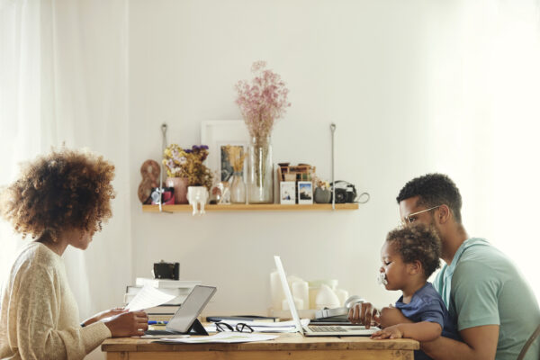 Mother using digital tablet for working from home. Father using laptop with son at table. They are sitting in kitchen.