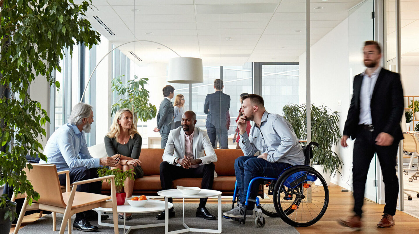 Business associates in 20s, 40s, and 50s relaxing and exchanging ideas in sitting area of modern office.