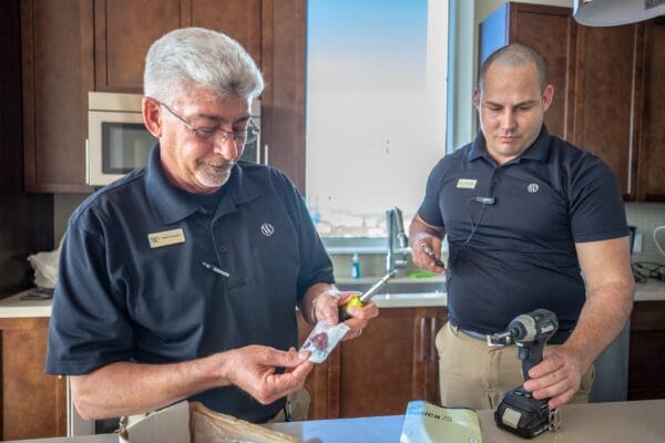 Two men in black shirts and pants collaborate in a kitchen, engaged in food preparation and cooking tasks.